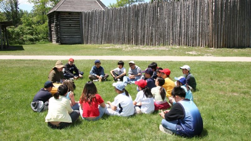A group of young visitors sits in a circle with a historical interpreter
