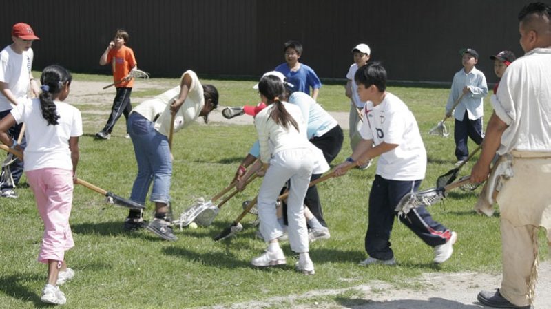A group of young visitors, supervised by an Indigenous interpreter, playing lacrosse