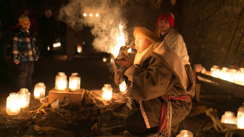 A costumed historical interpreter, illuminated by candles, demonstrates traditional firestarting techniques