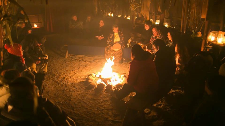 Des ainés autochtones et plusieurs visiteurs dans une maison longue à Sainte-Marie, illuminés par un feu et plusieurs bougies.