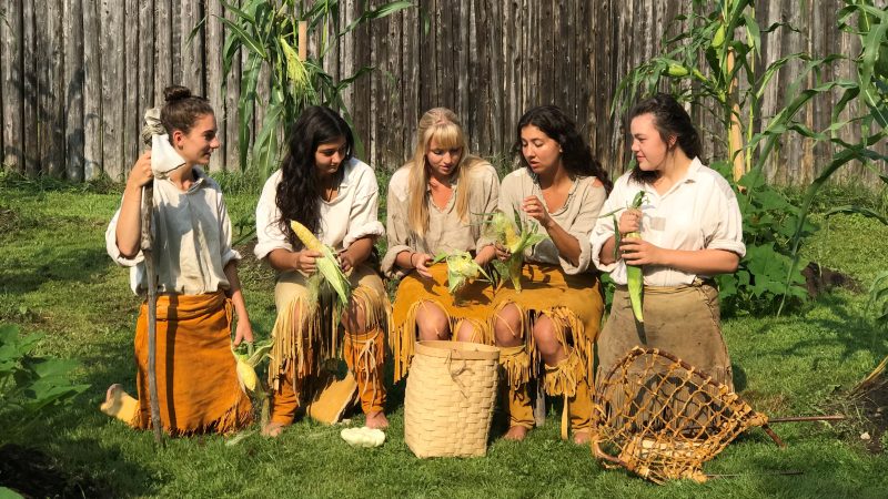Cinq interprètes historiques autochtones, en costume de Huron-Wendat, a éplucher du blé d'inde