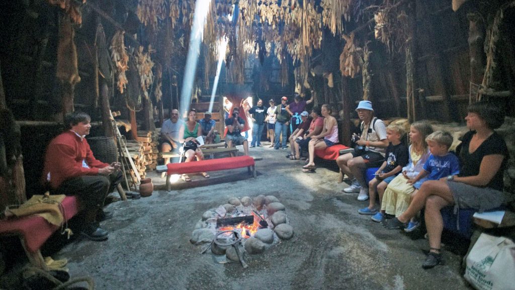 A group of visitors, of all ages, listens to stories told by a costumed interpreter in the longhouse at Sainte-Marie among the Hurons