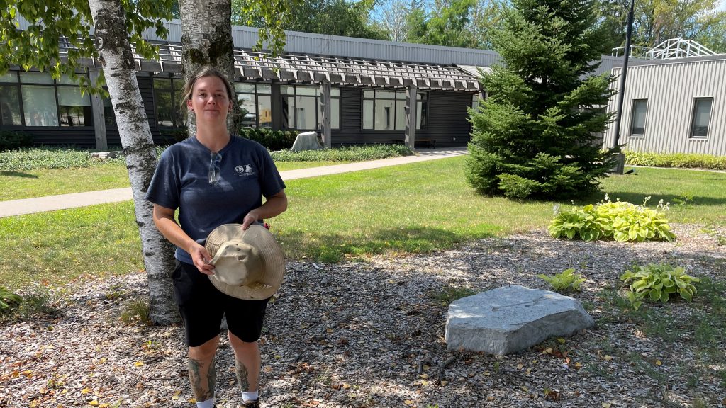 Katie, wearing safety glasses and a Sainte-Marie among the Hurons T-shirt, stands in front of the administration centre of Huronia Historical Parks. Katie is member of our maintenance crew for 2022.