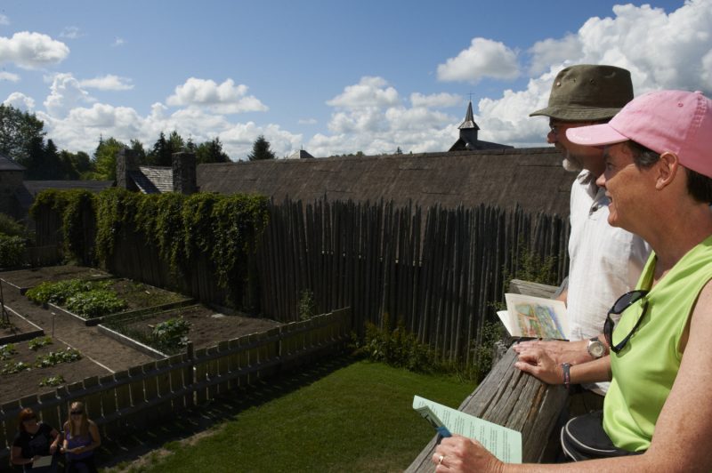 Two visitors stand on the palisade walls, holding maps, and look down at the historic site