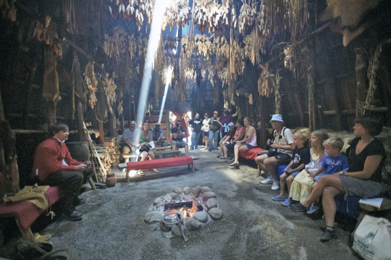 A group of visitors, of all ages, listens to stories told by a costumed interpreter in the longhouse at Sainte-Marie among the Hurons