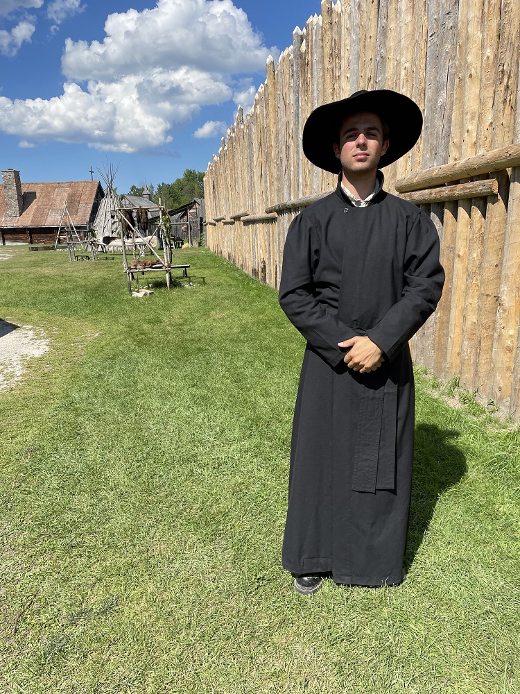 A historical interpreter in a jesuit cassock standing by the palisade walls at Sainte-Marie among the Hurons