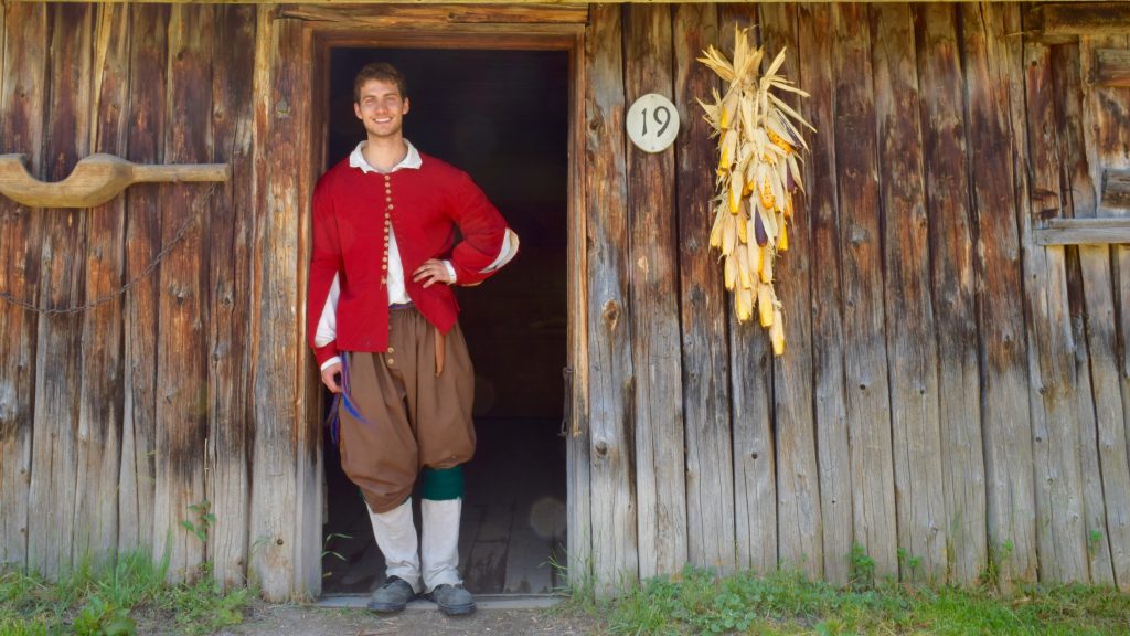 A costumed historical interpreter leans against a door jam, smiling