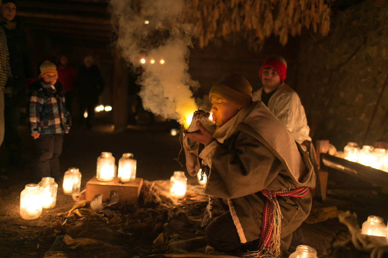 Une homme métis qui démontre la méthode traditionelle autochtones de commencer un feu. Il souffle sur un braise dans un morceau d'écorce de bouleau. C'est la nuit et il en entouré de chandelles en crocs.