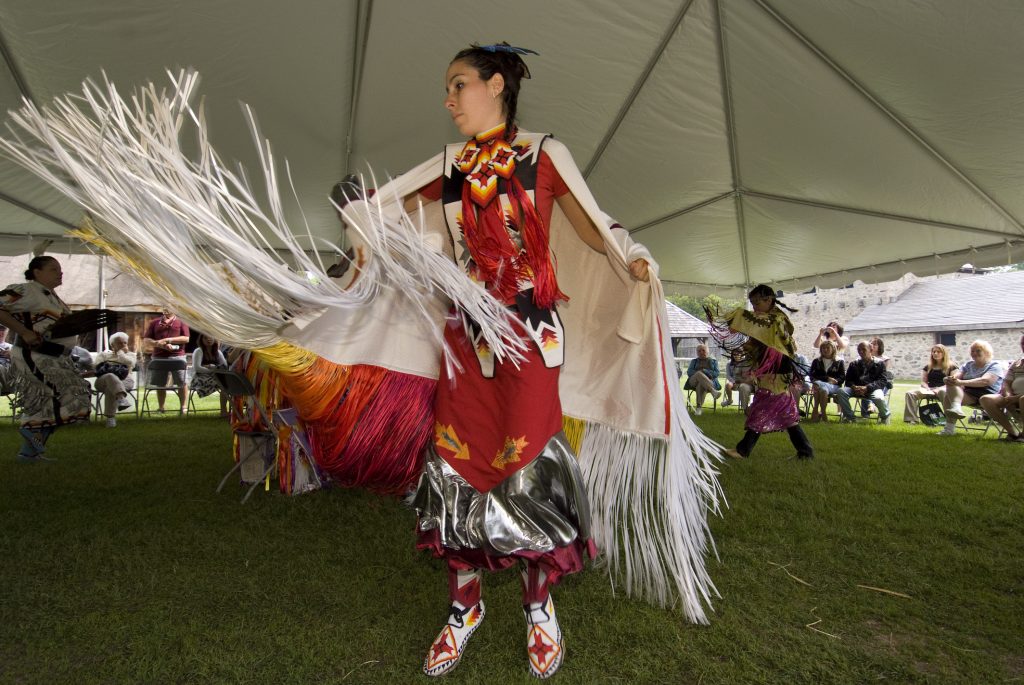 A First Nations woman in ceremonial dress dancing under a tent a Sainte-Marie