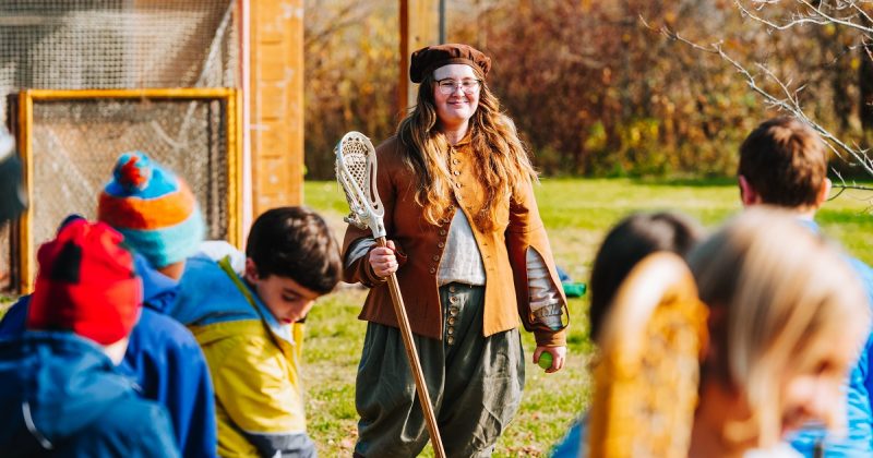 Hope, a smiling interpreter in costume, holding a lacross stick and watching students as they practise their lacrosse skills