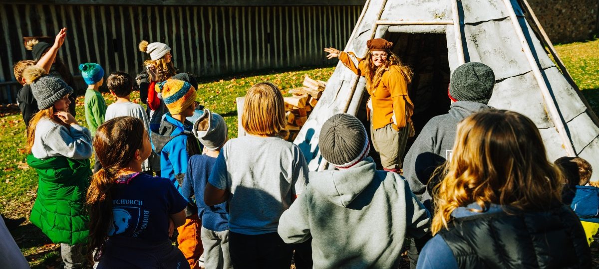 Hope, a costumed interpreter at Sainte-marie, entertaining a group of students outside of the main entrance wigwam