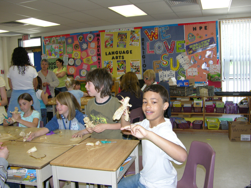 A classroom with students making cornhusk doll. One smiling student holds up his doll.