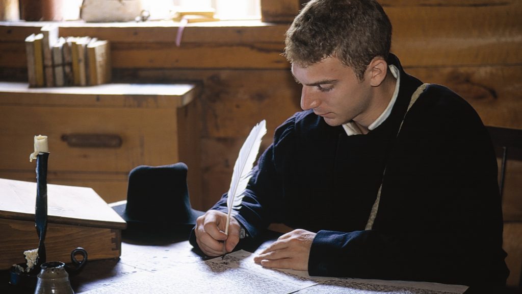 Costumed interpreter wearing a Jesuit cassock, writing a letter with a quill pen