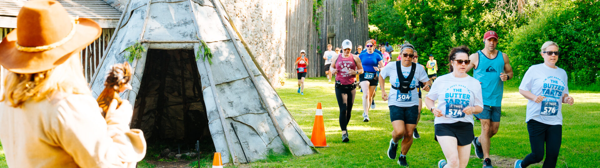 Racers running past a wigwam on the grounds at Sainte-Marie